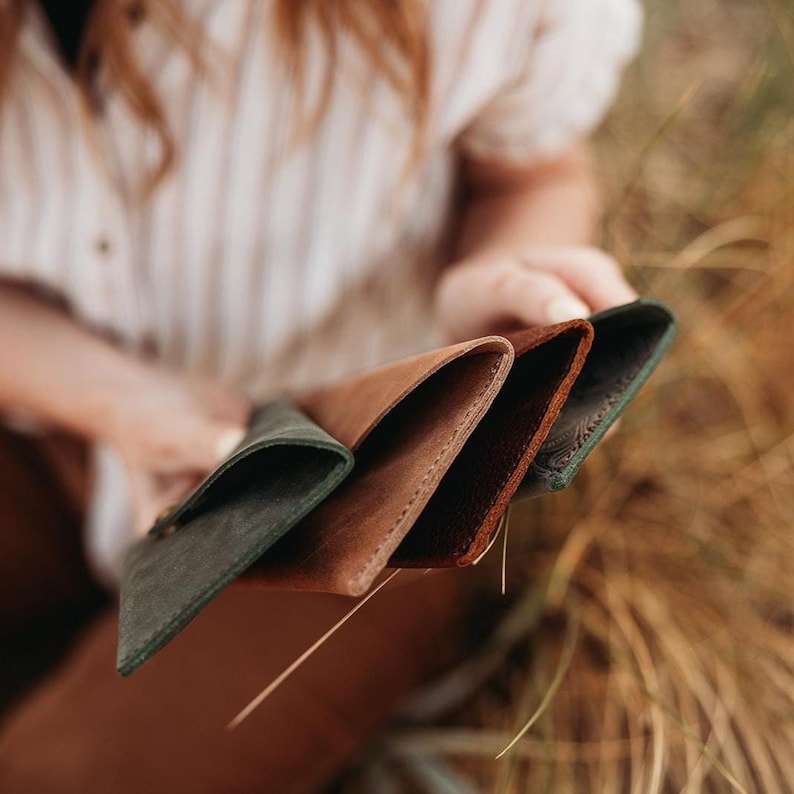 May include: A selection of leather wallets in various colors, including dark green, brown, and reddish-brown. The wallets are held in a person's hands, showcasing their different textures and hues. The wallets appear to be new and unused.