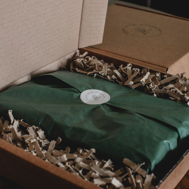 May include: A cardboard box with a dark green wrapped package inside, secured with a white sticker. The box is filled with brown paper filler. The sticker has the text "Ruth Handmade" in a circular design.