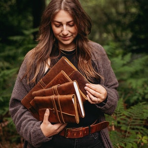 Könnte beinhalten: Eine Frau hält einen Stapel von vier braunen, ledergebundenen Tagebüchern mit Riemen. Die Tagebücher sind in einem Stapel gestapelt, wobei das oberste Tagebuch das größte und das unterste Tagebuch das kleinste ist. Die Tagebücher sind alle geschlossen und haben ein rustikales, handgemachtes Aussehen.