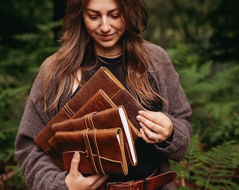 May include: A woman holds a stack of four brown leather-bound journals with straps. The journals are stacked in a pile, with the top journal being the largest and the bottom journal being the smallest. The journals are all closed and have a rustic, handmade look.