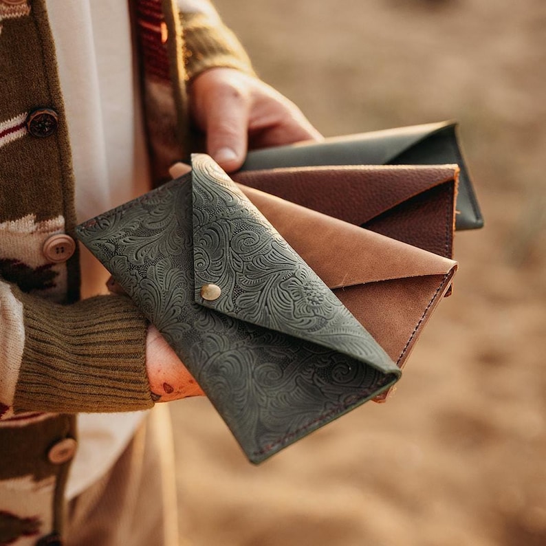 May include: A collection of leather eyeglass cases in various colors. The front-most case is dark green with an embossed floral pattern and a brass snap closure. Other cases are brown and tan.