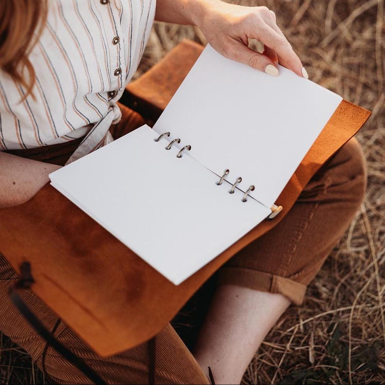 May include: Open leather journal with blank white pages. The journal is brown with a ring-bound spine. The person is wearing a striped shirt and brown pants. The journal is open, revealing the blank pages.