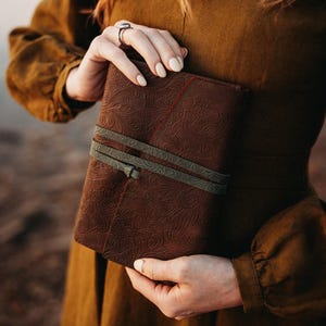 May include: A brown leather journal with an embossed floral pattern is held. The journal is secured with two olive green straps. The journal's cover has a detailed, textured design. The person is wearing a brown dress.