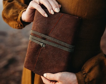 May include: A brown leather journal with an embossed floral pattern is held by a person. The journal is secured with two olive green straps. The journal's cover has a detailed, textured design. The person is wearing a brown dress.