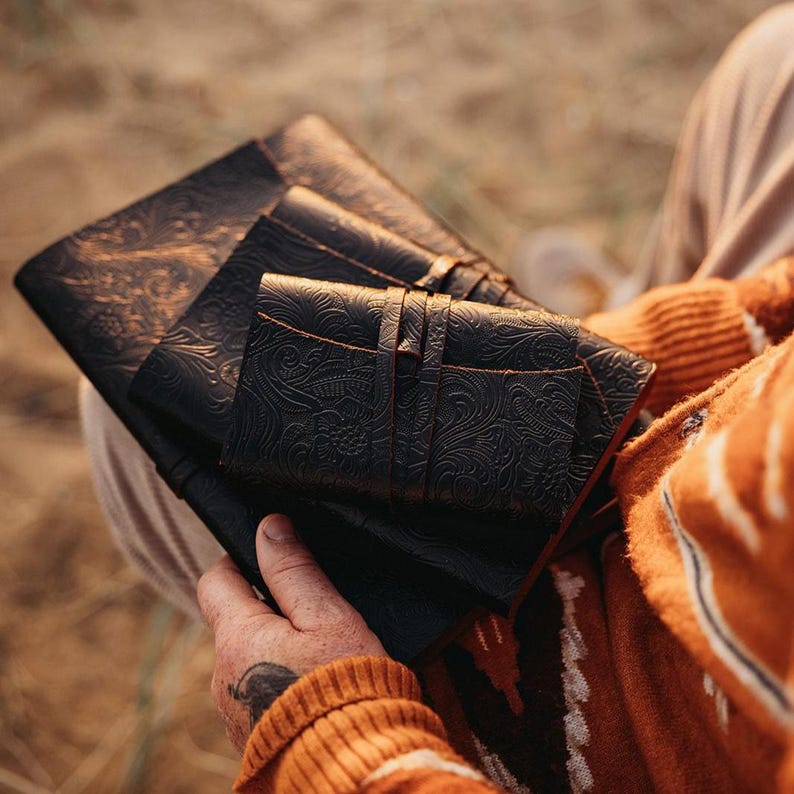 May include: A stack of three black leather journals with embossed floral patterns. The journals are of varying sizes, with the smallest one secured by a leather strap. The leather has a rich, dark color, and the edges are slightly distressed.