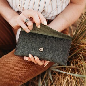 May include: Dark green leather wallet with an embossed floral pattern. The wallet has a snap closure and a contrasting red stitch. The interior is a lighter shade of green. The wallet is open, revealing the inside.