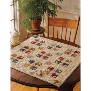 May include: A quilted table runner with a repeating pattern of colorful teacups. The teacups are in shades of red, blue, brown, and purple, set against a cream background. A pair of scissors and a spool of thread are on the table.