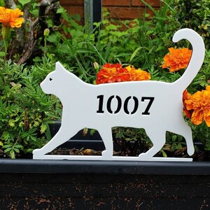 May include: A white cat-shaped house number sign with the number 1007 in black. The sign is placed in front of a vibrant display of orange marigold flowers and green foliage.