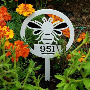 May include: White metal garden stake featuring a bee design within a circular frame. The stake displays the number "951" below the bee. The stake is placed in a garden with orange and yellow flowers and green foliage.