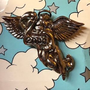 May include: A silver angel pin with wings outstretched, holding a banner with the year 2000 printed on it. The pin is on a blue and white background with clouds and stars.