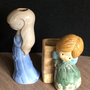 May include: Two ceramic figurines of girls, one with blonde hair wearing a blue dress and the other with brown hair wearing a green dress. Both figurines are holding flowers and have a vintage style.