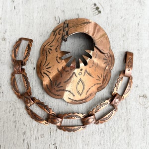 May include: A copper necklace with a large, ornate pendant. The pendant is shaped like a flower and has a cutout in the center. The necklace is made of linked copper pieces with intricate designs.