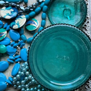 May include: Teal blue glass jar with a textured lid and the word "AVON" printed on the bottom. The jar is surrounded by blue and white beads.
