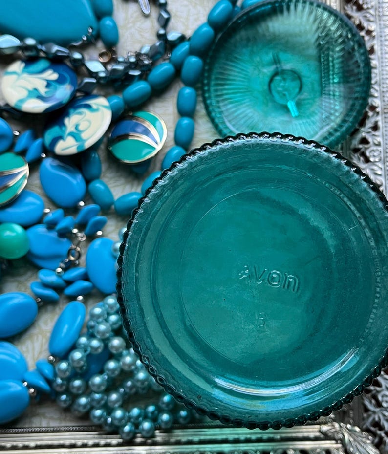 May include: Teal blue glass jar with a textured lid and the word "AVON" printed on the bottom. The jar is surrounded by blue and white beads.
