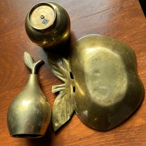 May include: A set of three brass fruit-shaped decorative bowls. The bowls are shaped like an apple, a pear, and a round apple. The bowls are all polished and have a shiny finish.