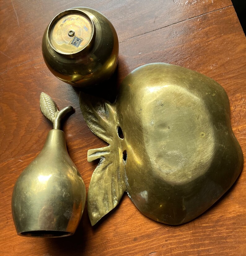 May include: A set of three brass fruit-shaped decorative bowls. The bowls are shaped like an apple, a pear, and a round apple. The bowls are all polished and have a shiny finish.
