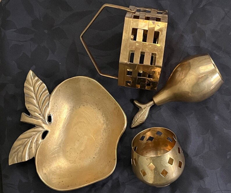 May include: A set of four gold-toned metal decorative items. The items include a small apple-shaped dish, a pear-shaped dish, a small basket with a handle, and a small round dish with a geometric pattern.