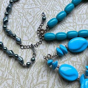 May include: Two necklaces, one with dark gray beads and a silver clasp, and the other with turquoise blue beads and a silver clasp.