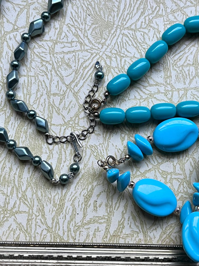 May include: Two necklaces, one with dark gray beads and a silver clasp, and the other with turquoise blue beads and a silver clasp.