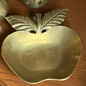 May include: A gold-toned metal apple-shaped dish with a leaf design. The dish is made of brass and has a smooth, polished finish.
