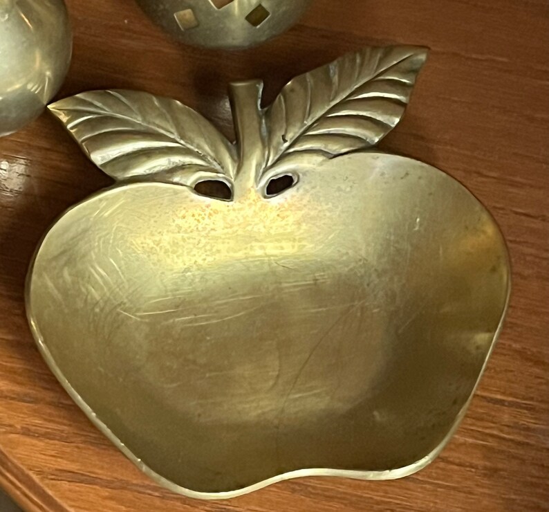May include: A gold-toned metal apple-shaped dish with a leaf design. The dish is made of brass and has a smooth, polished finish.