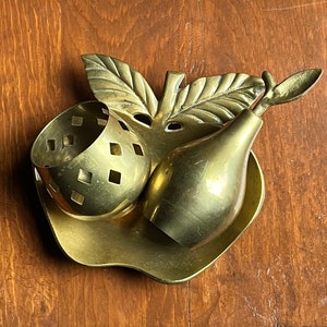 May include: A gold-toned metal trinket dish shaped like an apple with a pear and a small bowl with a geometric pattern. The dish is on a wooden surface.