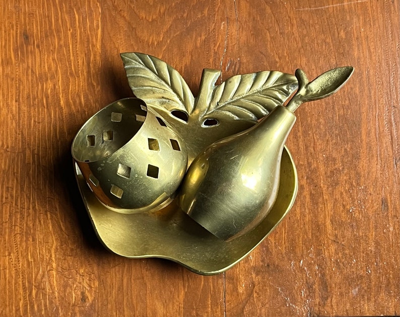 May include: A gold-toned metal trinket dish shaped like an apple with a pear and a small bowl with a geometric pattern. The dish is on a wooden surface.