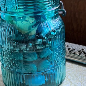 May include: A teal blue glass jar with a decorative pattern and a metal lid. The jar has a wire handle on each side. The jar is filled with various items, including flowers and other decorative elements.
