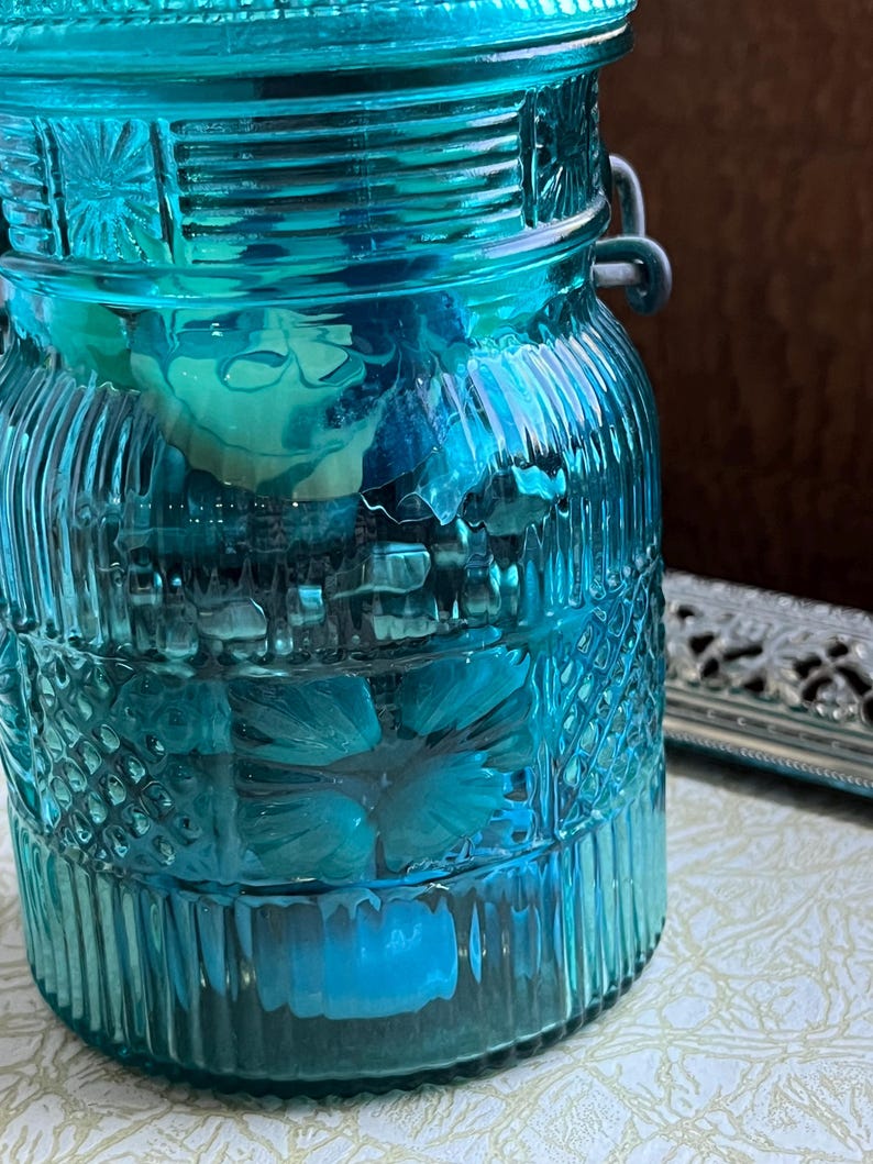 May include: A teal blue glass jar with a decorative pattern and a metal lid. The jar has a wire handle on each side. The jar is filled with various items, including flowers and other decorative elements.