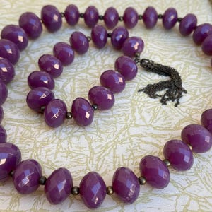 May include: A long purple beaded necklace with a silver chain tassel. The beads are faceted and have a glossy finish.
