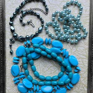 May include: Four strands of blue and teal colored beaded necklaces. The necklaces are made of various sizes and shapes of beads. The necklaces are displayed on a white background with a silver frame.