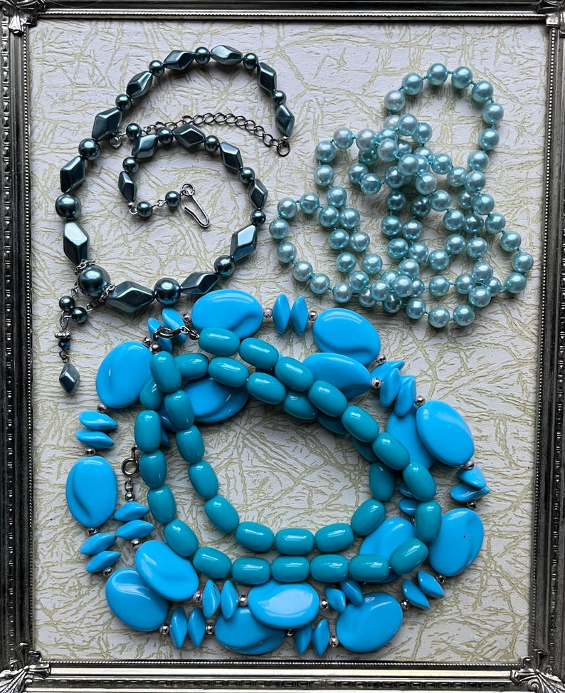 May include: Four strands of blue and teal colored beaded necklaces. The necklaces are made of various sizes and shapes of beads. The necklaces are displayed on a white background with a silver frame.
