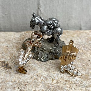 May include: A miniature silver horse figurine with a pack on its back, next to a gold shovel charm and a gold cowboy boot charm, both adorned with clear rhinestones. The charms are on a textured surface.