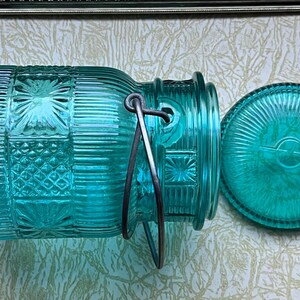 May include: A teal blue glass jar with a metal handle and a matching lid. The jar has a textured, patterned design.