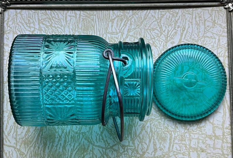 May include: A teal blue glass jar with a metal handle and a matching lid. The jar has a textured, patterned design.