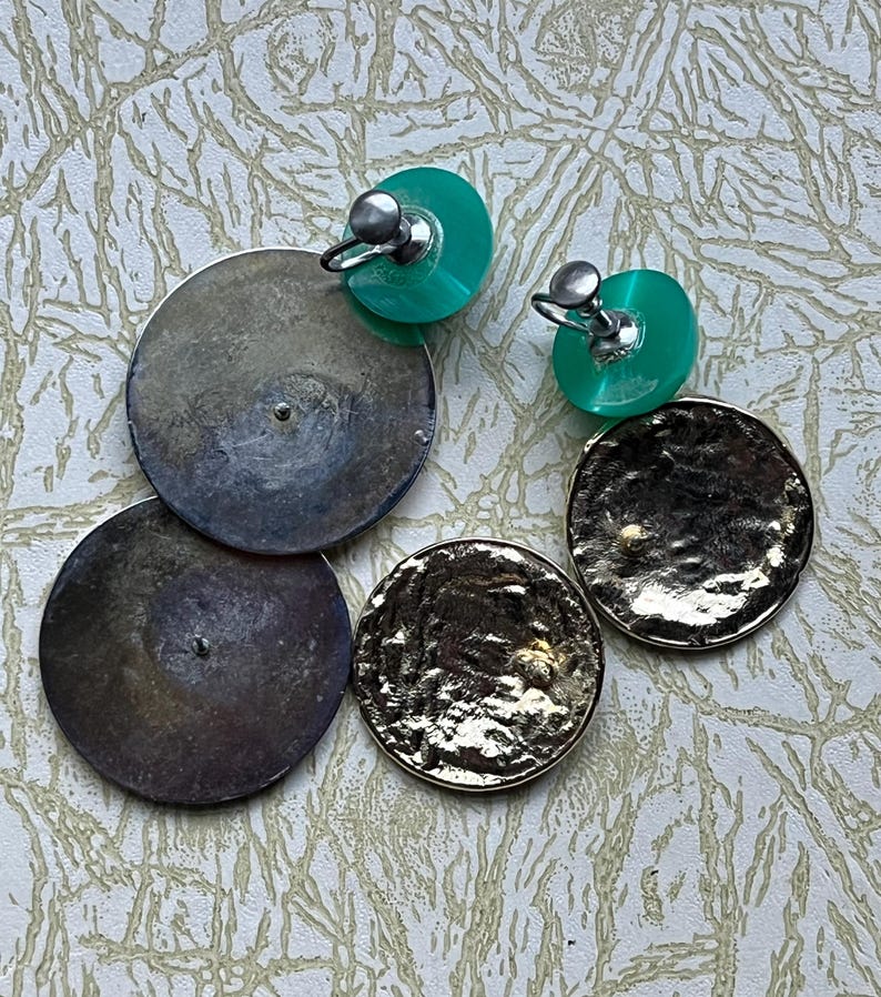 May include: A pair of clip-on earrings with two round discs. One disc is silver and the other is gold. The clip-on part of the earring is green.