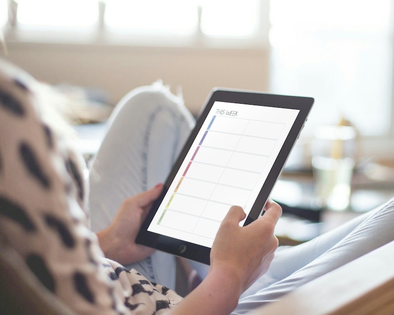 May include: A person is holding a tablet with a weekly planner on the screen. The planner has a rainbow coloured column on the left side and blank spaces for each day of the week.