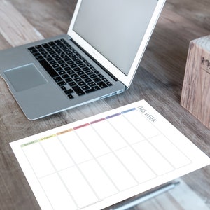 May include: A white weekly planner with a rainbow coloured header and the text "This Week". The planner is on a wooden table with a laptop computer and a wooden pencil holder.