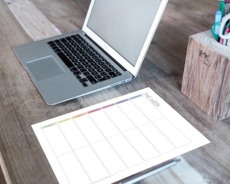 May include: A white weekly planner with a rainbow coloured header and the text "This Week". The planner is on a wooden table with a laptop computer and a wooden pencil holder.