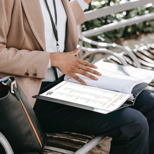 May include: A person wearing a beige blazer and black pants sits on a bench and looks at a black binder with a white page open. The binder has the text "Gift Registry" on the front.