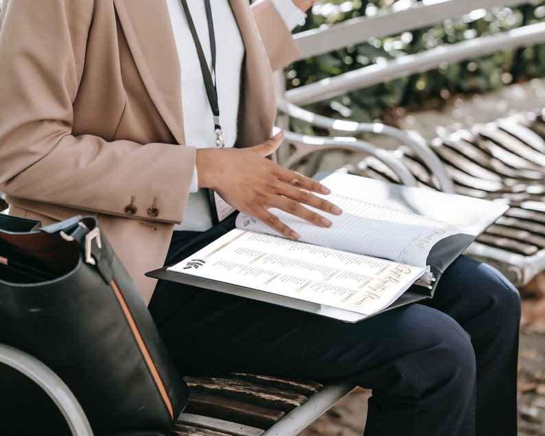 May include: A person wearing a beige blazer and black pants sits on a bench and looks at a black binder with a white page open. The binder has the text "Gift Registry" on the front.