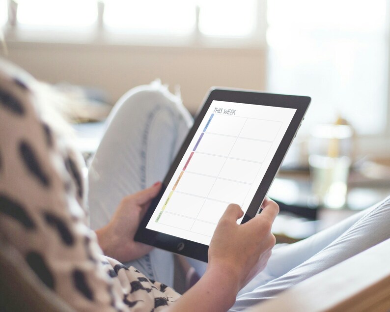 May include: A person is using a tablet to plan their week. The tablet screen shows a weekly planner with the words "This Week" at the top. The planner is divided into days of the week and has space for writing tasks or appointments.