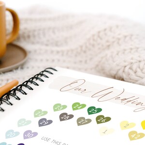 May include: A notebook with a spiral binding and a white cover. The cover has the text "Our Wedding" written in brown cursive. The notebook is open to a page with a color chart of small hearts with numbers on them. The text "USE THIS" is partially visible at the bottom of the page.