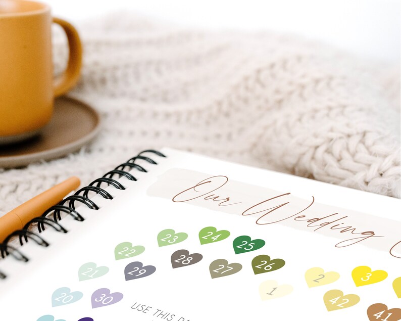 May include: A notebook with a spiral binding and a white cover. The cover has the text "Our Wedding" written in brown cursive. The notebook is open to a page with a color chart of small hearts with numbers on them. The text "USE THIS" is partially visible at the bottom of the page.