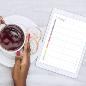 May include: A white tablet with a weekly planner on the screen. The planner has a colourful column for each day of the week. The tablet is on a white wooden surface with a cup of tea and biscuits.
