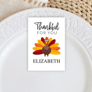 May include: A white rectangular place card with the text "Thankful FOR YOU" and a cartoon turkey illustration. The turkey has a brown body, red, orange, and yellow feathers, and the name "ELIZABETH" is printed below the turkey.