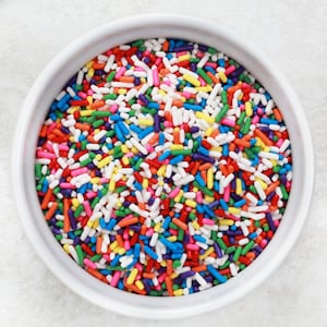May include: A white bowl filled with colorful sprinkles. The sprinkles are a mix of red, orange, yellow, green, blue, purple, and white.