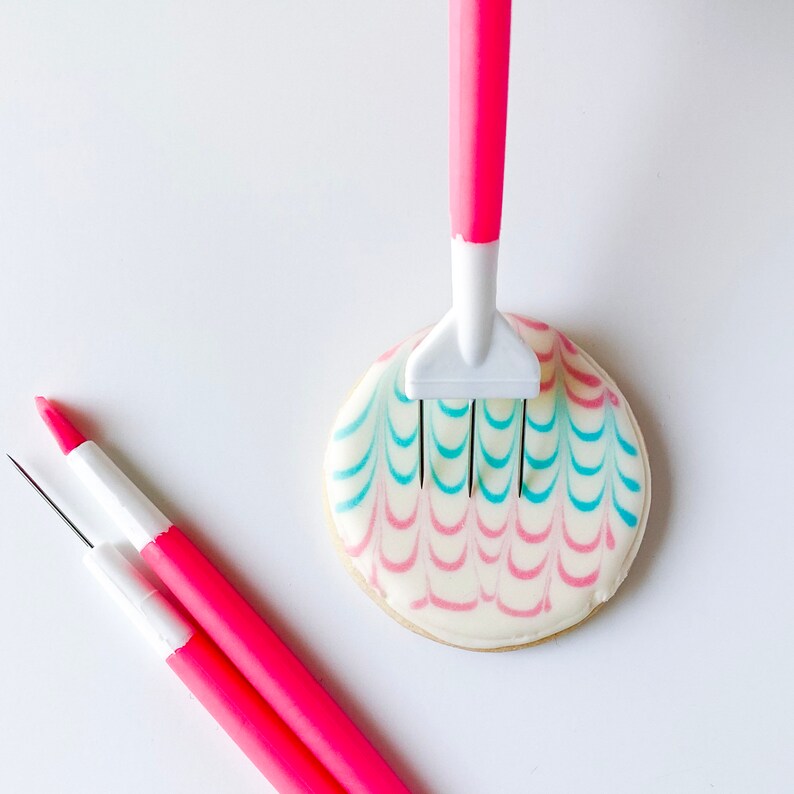 May include: A close-up of a pink and blue decorated cookie with a white icing comb tool being used to create a pattern in the icing.
