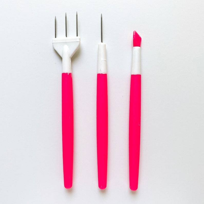 May include: Three pink and white tools for crafting. The tool on the left has three prongs, the middle tool has a single point, and the tool on the right has a rounded tip.