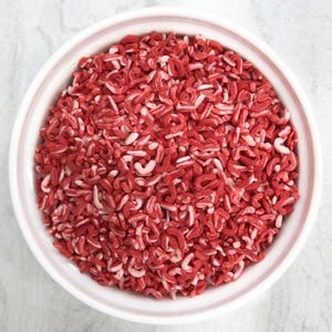 May include: A white ceramic bowl filled with red and white candy sprinkles. The sprinkles are small, curved shapes, and the bowl is set against a light-colored background. The candy is likely for decorating baked goods.